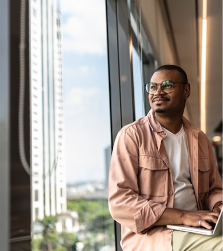 Man sits with laptop, gazing out sunny office window, wearing glasses and casual attire.