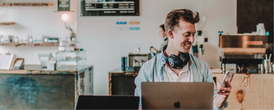 Young man with headphones uses phone and laptop in cozy cafe setting.